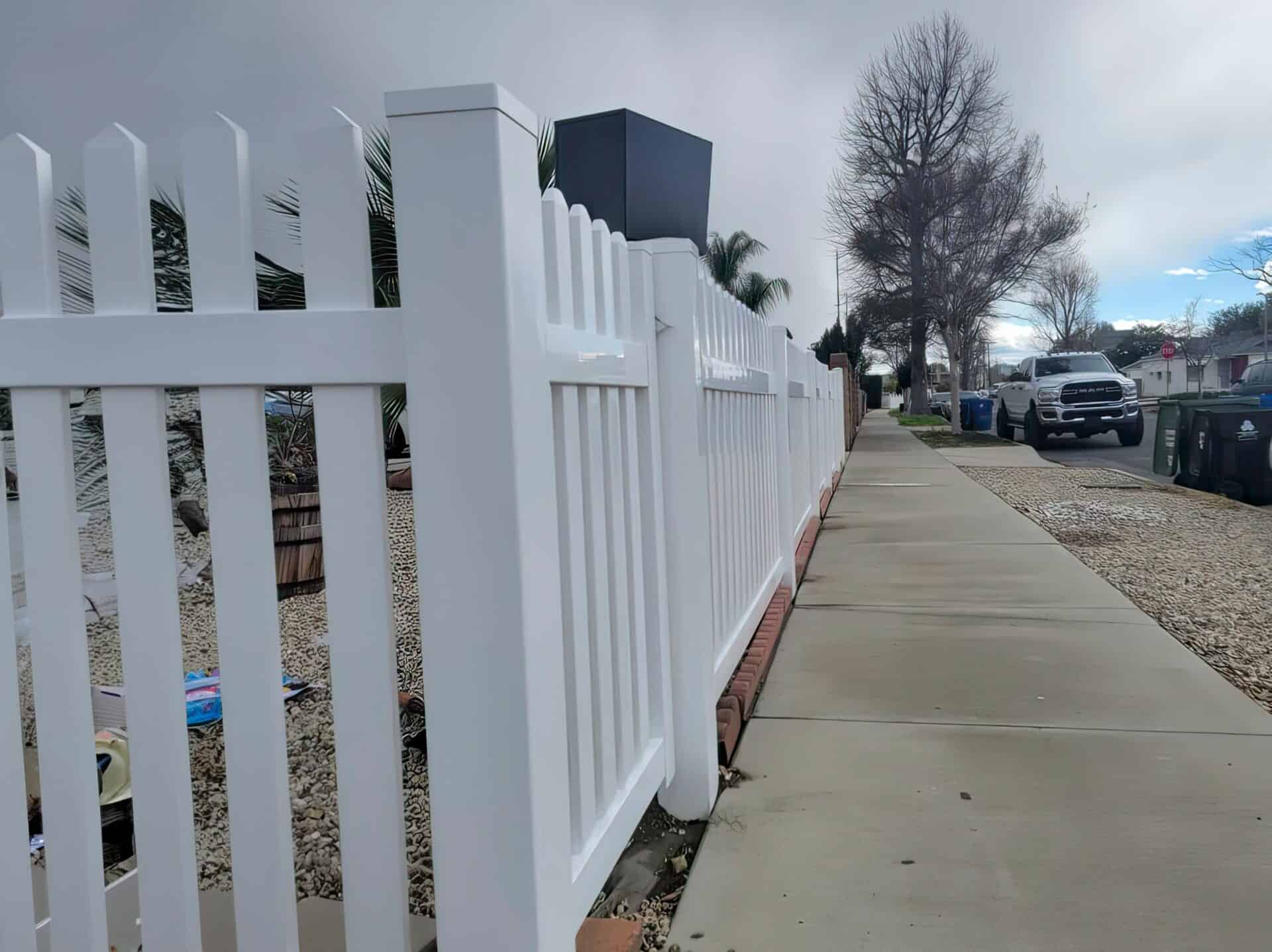A White Vinyl Vertical Picket Fence lines the footpath alongside tall trees and a parked car.