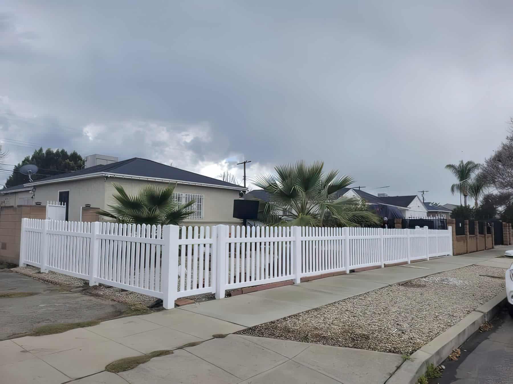 Vinyl vertical picket fence surrounding the house with tall palm trees in the front yard and a pavement outside it.